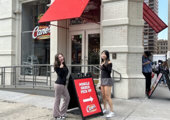 Zamira Frost and Miranda Chao Hwang standing in front of Raising Cane's, a brick restaurant with a red awning on a city corner.